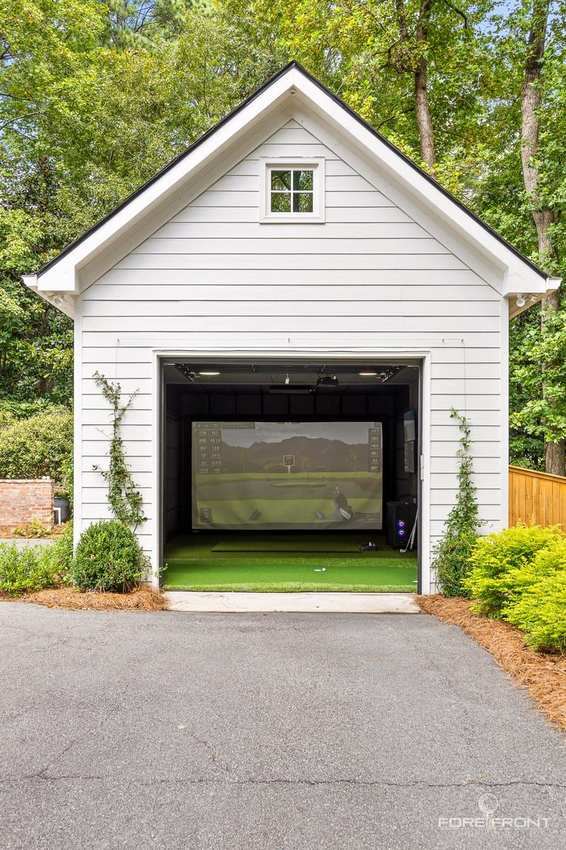 A detached garden building converted to a golf simulator bay, seen from the driveway with the door open.