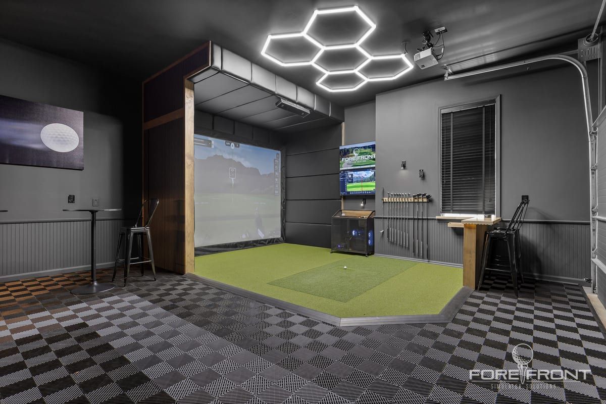 A converted garage golf simulator bay with charcoal walls, dark tiled flooring, hexagonal ceiling lights and an impact screen above short turf