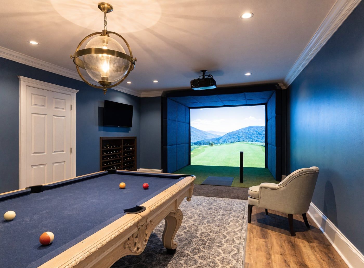A finished basement games room built around a golf simulator: impact screen in a padded blue enclosure showing a mountain fairway, a turf hitting area with a mat, a ceiling-mounted projector, and a pool table in the foreground